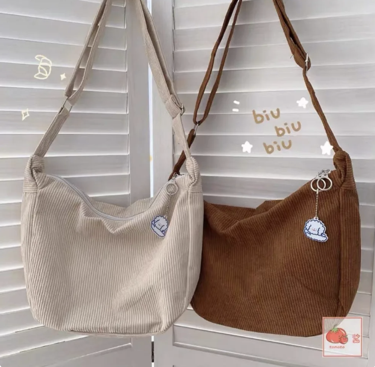 Dos bolsos de tela colgados en un perchero frente a persianas. Uno es beige y el otro marrón, ambos con un bordado de un puño. Fondo con texto "biu biu biu"