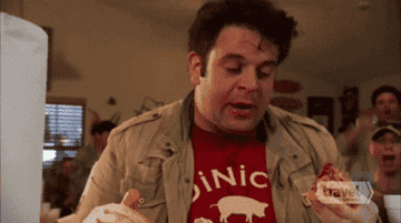 Adam Richman excitedly eats food, surrounded by cheering people in a casual dining setting