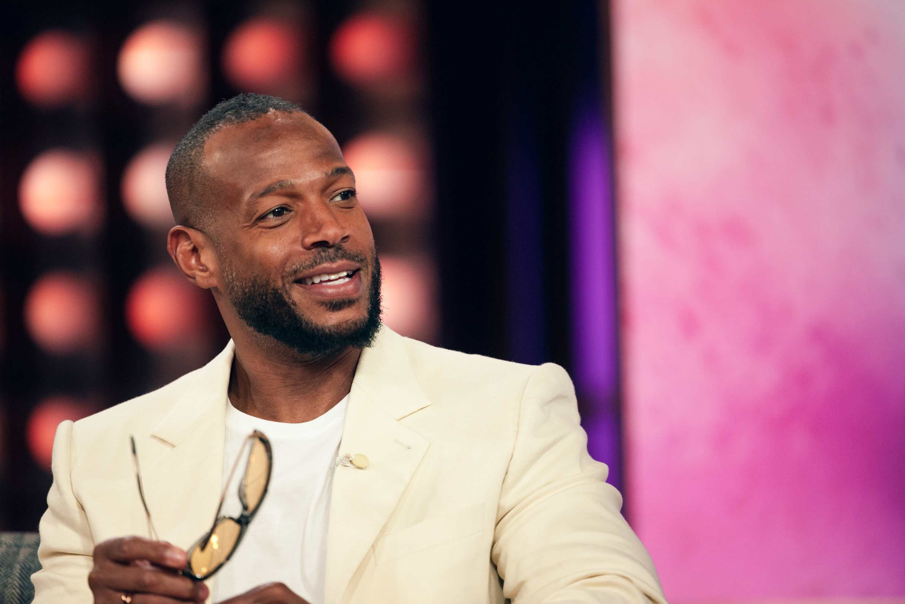 Marlon Wayans holds sunglasses and smiles while seated in a cream-colored suit jacket and white shirt, in an indoor setting