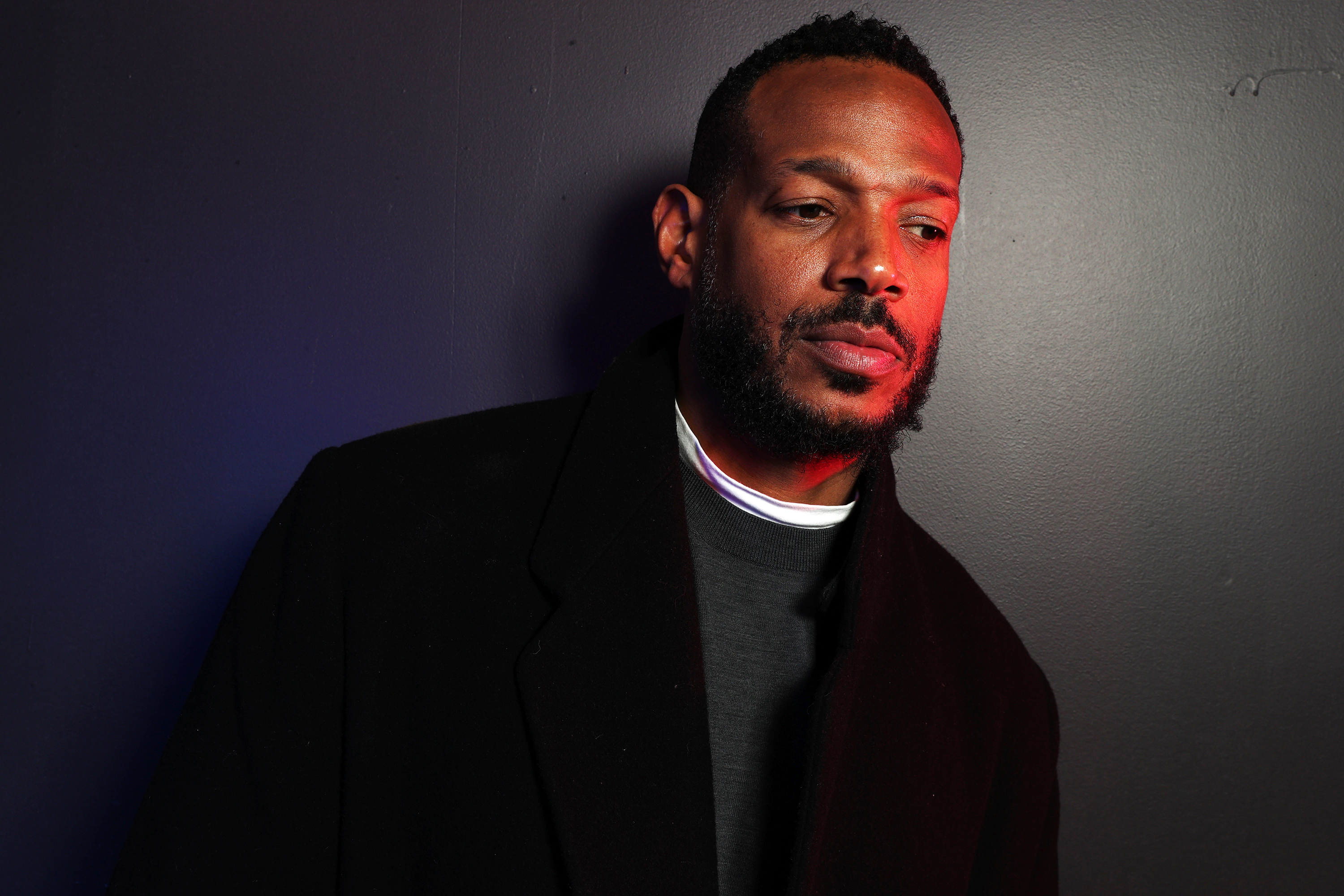 Marlon Wayans posing against a wall, dressed in a dark overcoat with a visible white shirt collar