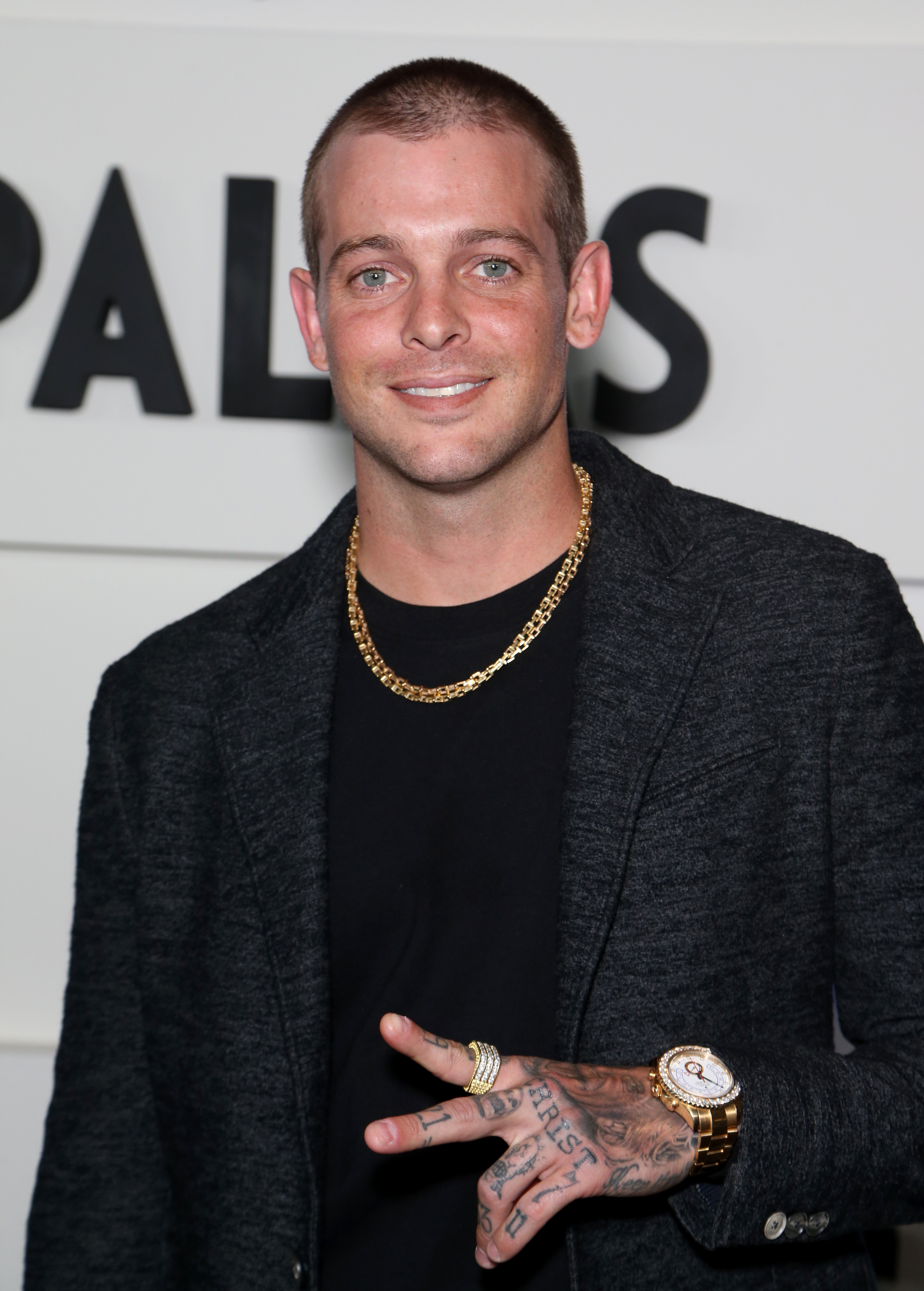 Ryan Sheckler with buzzed hair in a black shirt and gray blazer, wears a gold chain, gold watch, and ring. He is making a peace sign with tattooed fingers at an event.