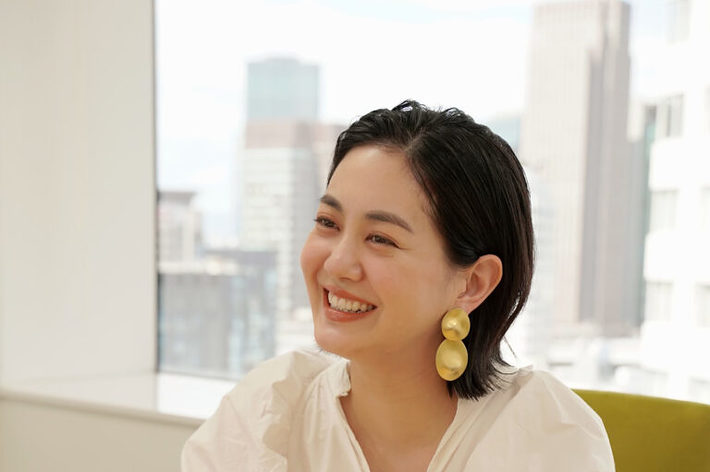女性がテーブルに座って微笑んでいます。白いブラウスと大きなイヤリングを身につけています。背景は都会のビル群。