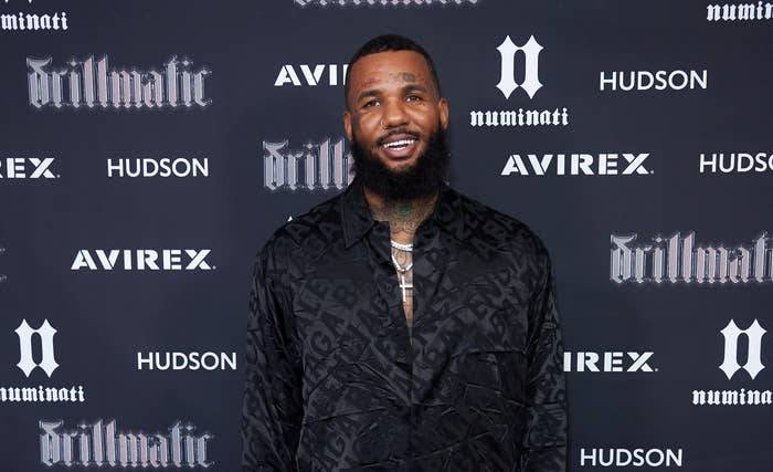 The Game poses on the red carpet in a black patterned suit at a music event, smiling. Logos of Avirex, Hudson, and &quot;drillmatic&quot; are visible in the background