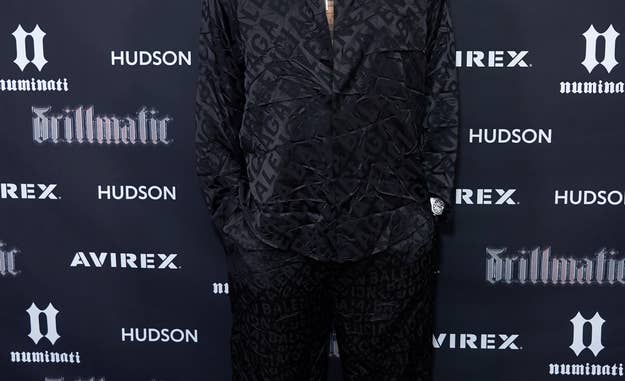 The Game poses on the red carpet in a black patterned suit at a music event, smiling. Logos of Avirex, Hudson, and "drillmatic" are visible in the background