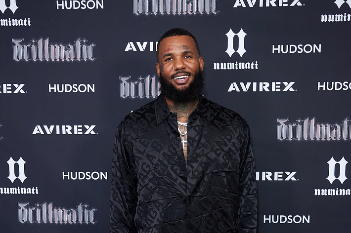 The Game poses on the red carpet in a black patterned suit at a music event, smiling. Logos of Avirex, Hudson, and "drillmatic" are visible in the background