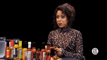 Liza Koshy is seated at a table with various hot sauces, eating chicken wings on the show &quot;Hot Ones.&quot;