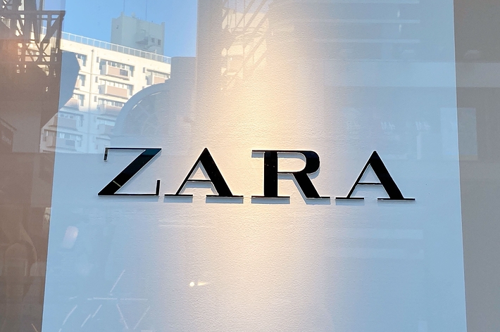 Storefront with large &ldquo;ZARA&rdquo; sign displayed on a transparent glass window. Urban buildings are reflected in the glass