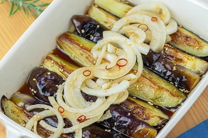 焼きナスの上に玉ねぎと唐辛子の輪切りがのった料理が白い陶器の容器に入っている。