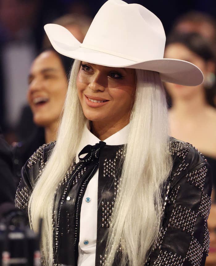 Beyonce is wearing a cowboy hat, a studded leather jacket, and a shirt with a bolo tie at an event. People are blurred in the background