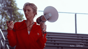 Jane Lynch in a sports tracksuit holding a megaphone and a stopwatch at a bleachers setting, featured in a Hulu and Disney+ content