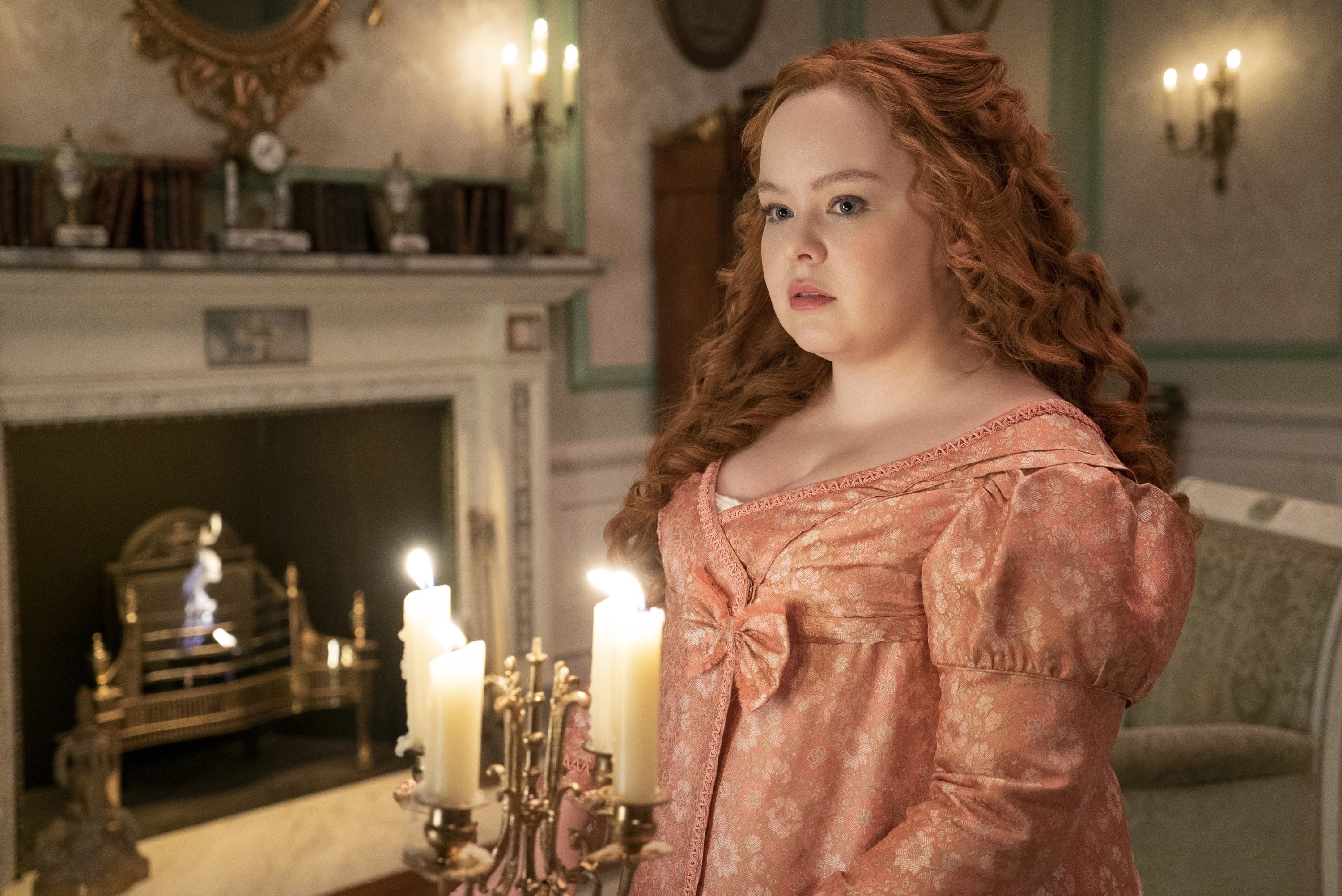 Nicola Coughlan stands in an elegant room wearing an ornate period dress, looking to the side. Candles and a fireplace are in the background