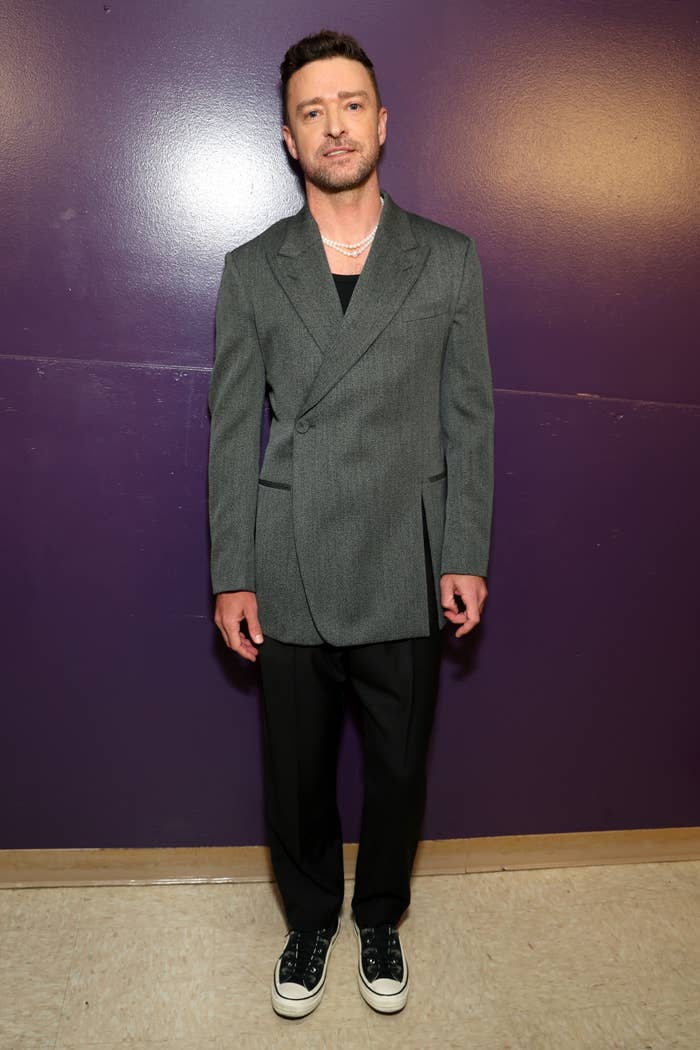 Justin Timberlake in a double-breasted blazer, black pants, and sneakers, standing against a plain wall