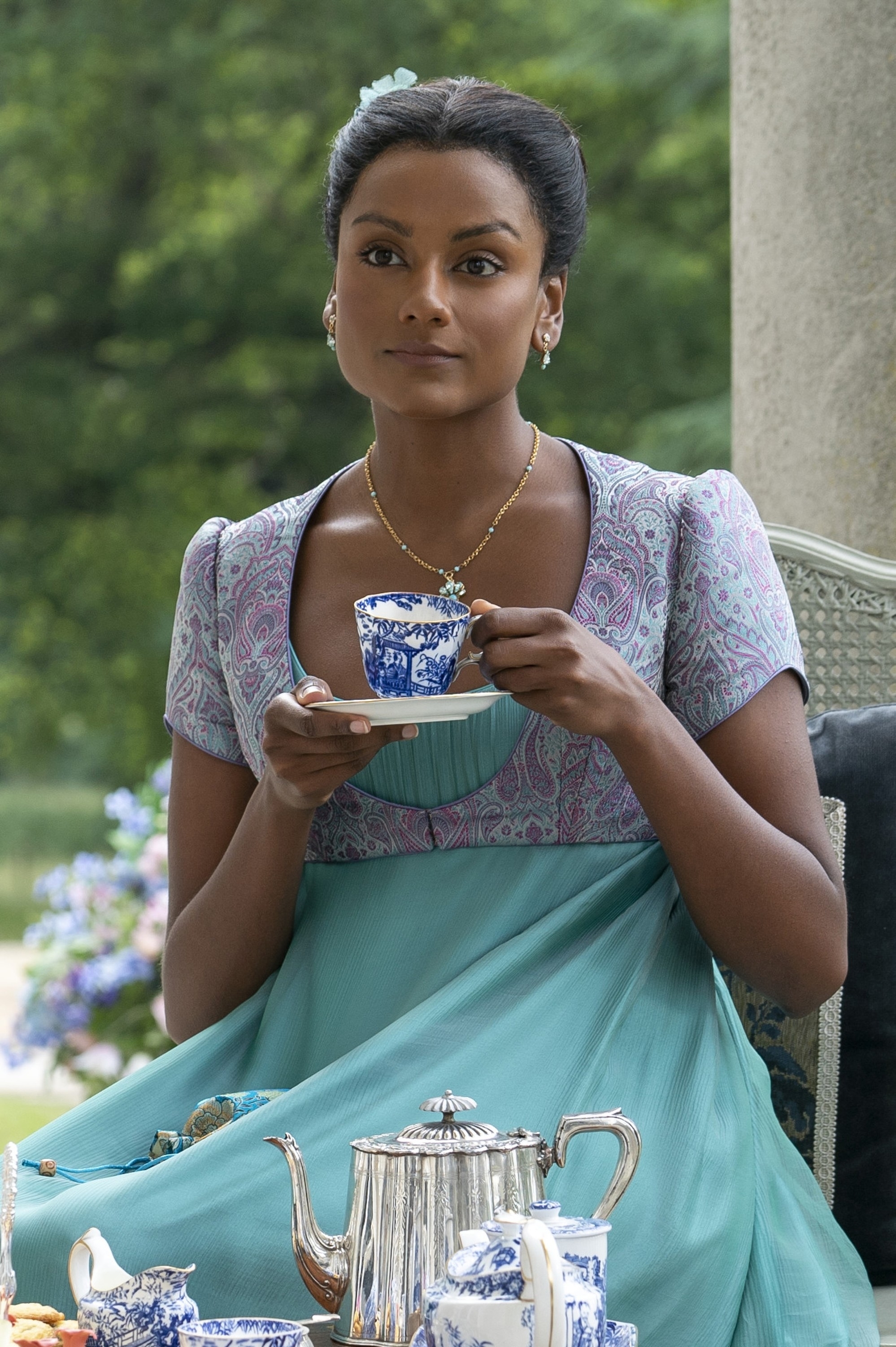 Simone Ashley dressed in Regency-era attire, holding a teacup and saucer, seated outdoors, with a teapot and other tea items on the table