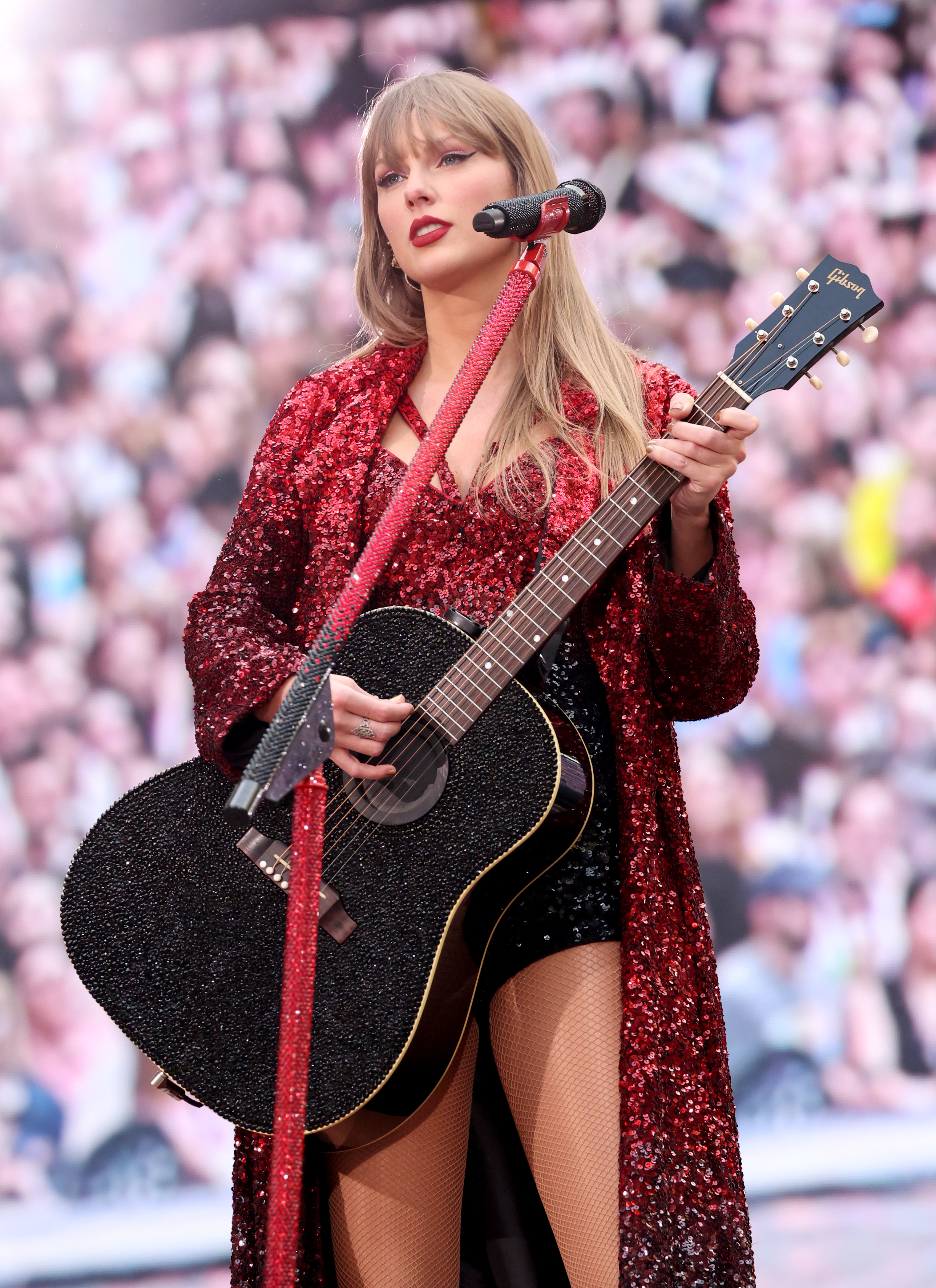 Taylor Swift performs on stage with a black guitar, wearing a glittery red jacket over a black outfit. The crowd is visible in the background