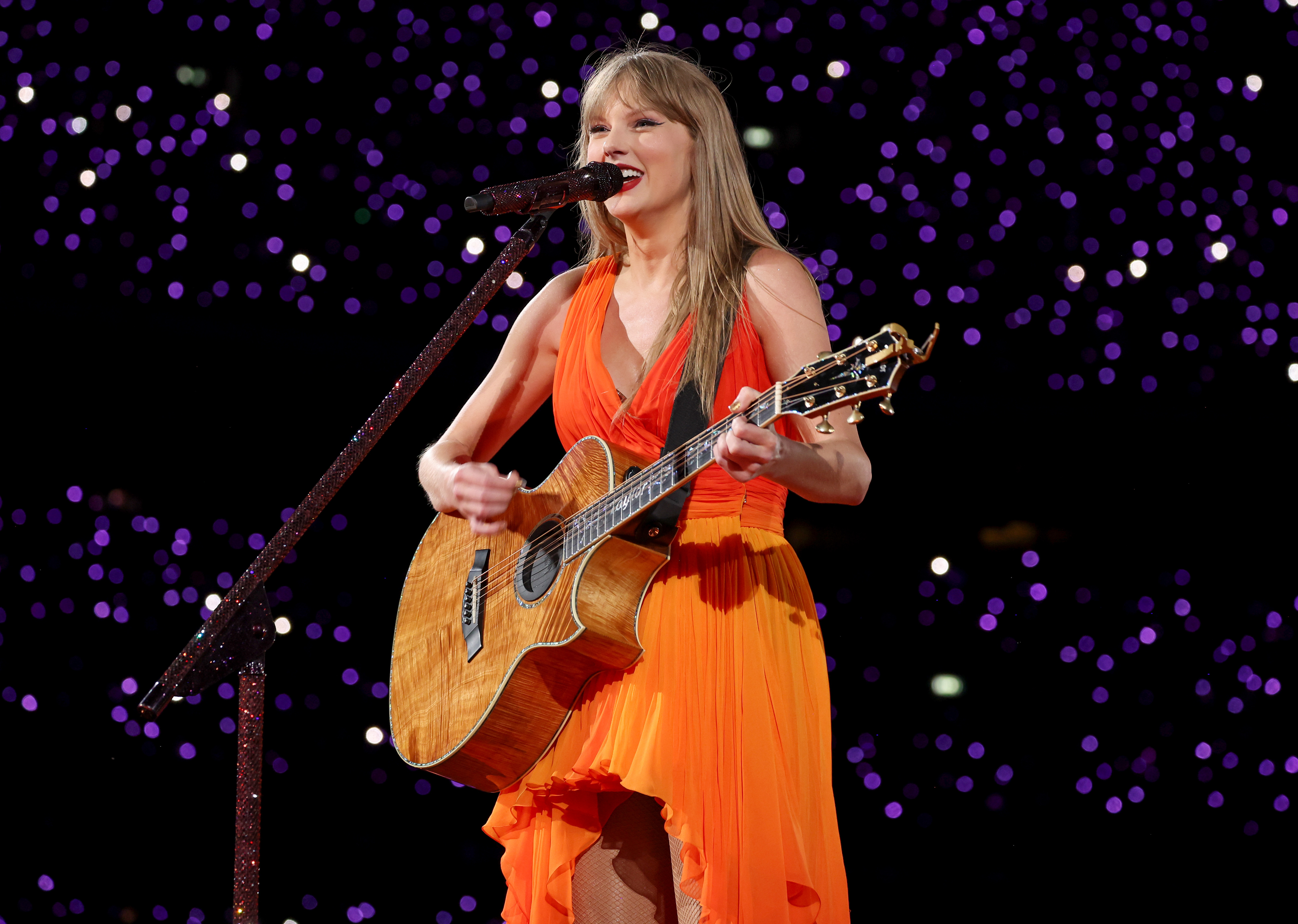 Taylor Swift performs on stage with a guitar, wearing a flowing, asymmetrical dress. Purple lights create a star-like background