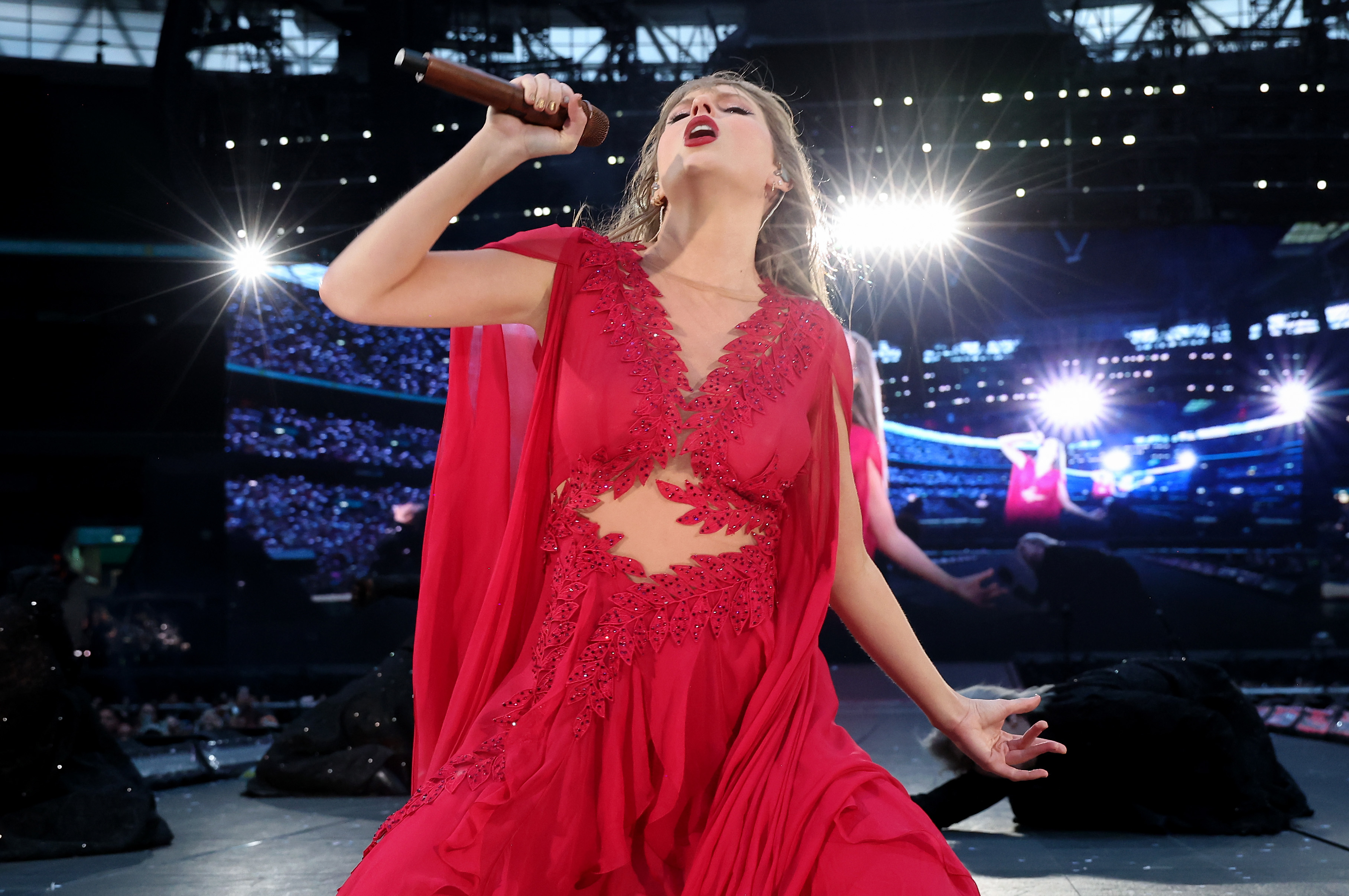 Taylor Swift performs passionately on stage, holding a microphone, wearing a flowing red dress with intricate detailing, against a backdrop of concert lights