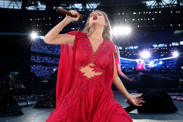 Taylor Swift performs passionately on stage, holding a microphone, wearing a flowing red dress with intricate detailing, against a backdrop of concert lights