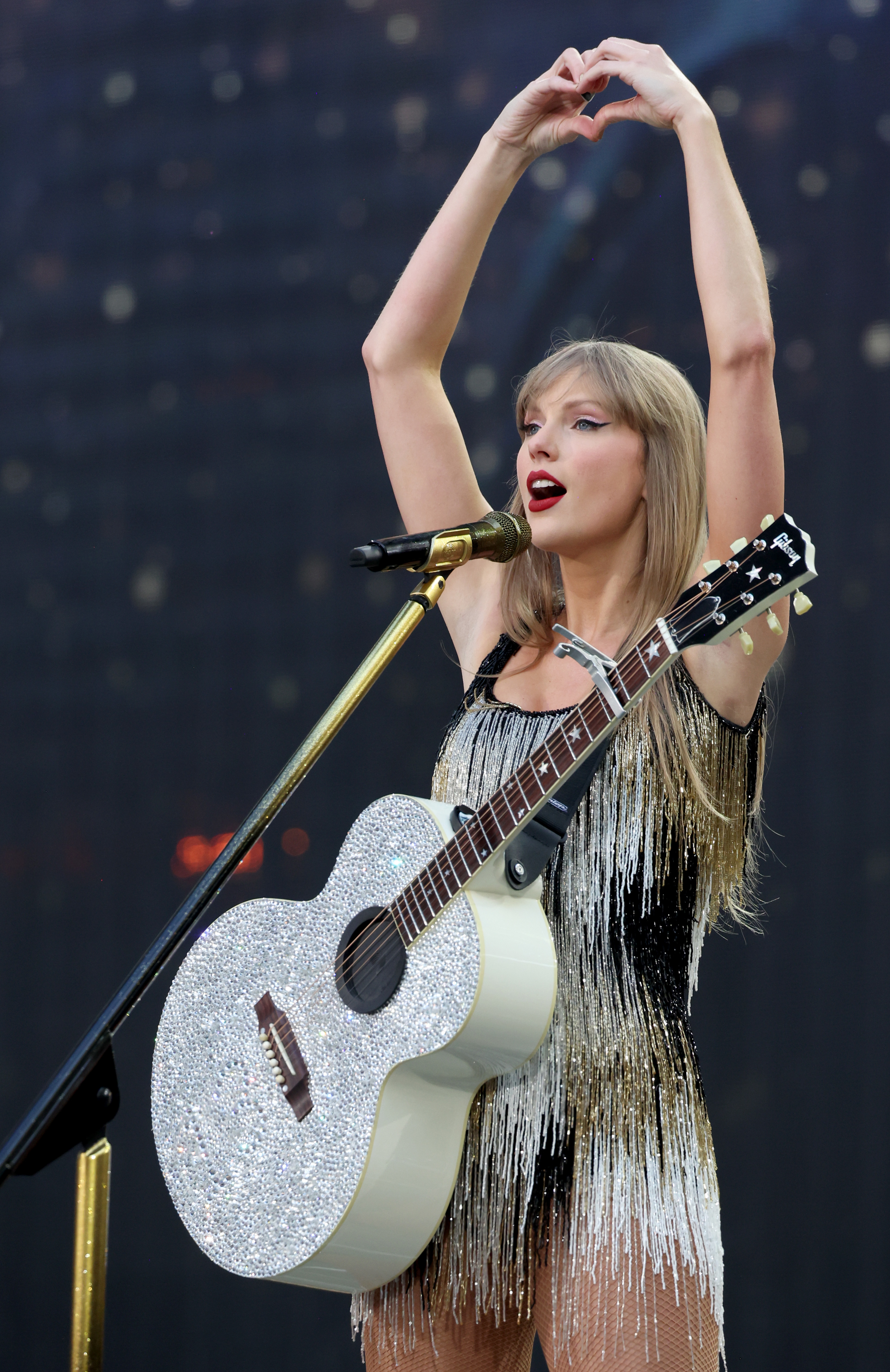 Taylor Swift performs on stage holding a sparkly white guitar, wearing a fringed outfit, and making a heart shape with her hands above her head