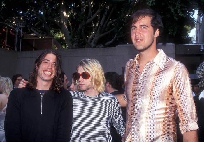 Dave Grohl, Kurt Cobain in a striped shirt, and Krist Novoselic in a polka-dot shirt at an outdoor event, smiling and posing together
