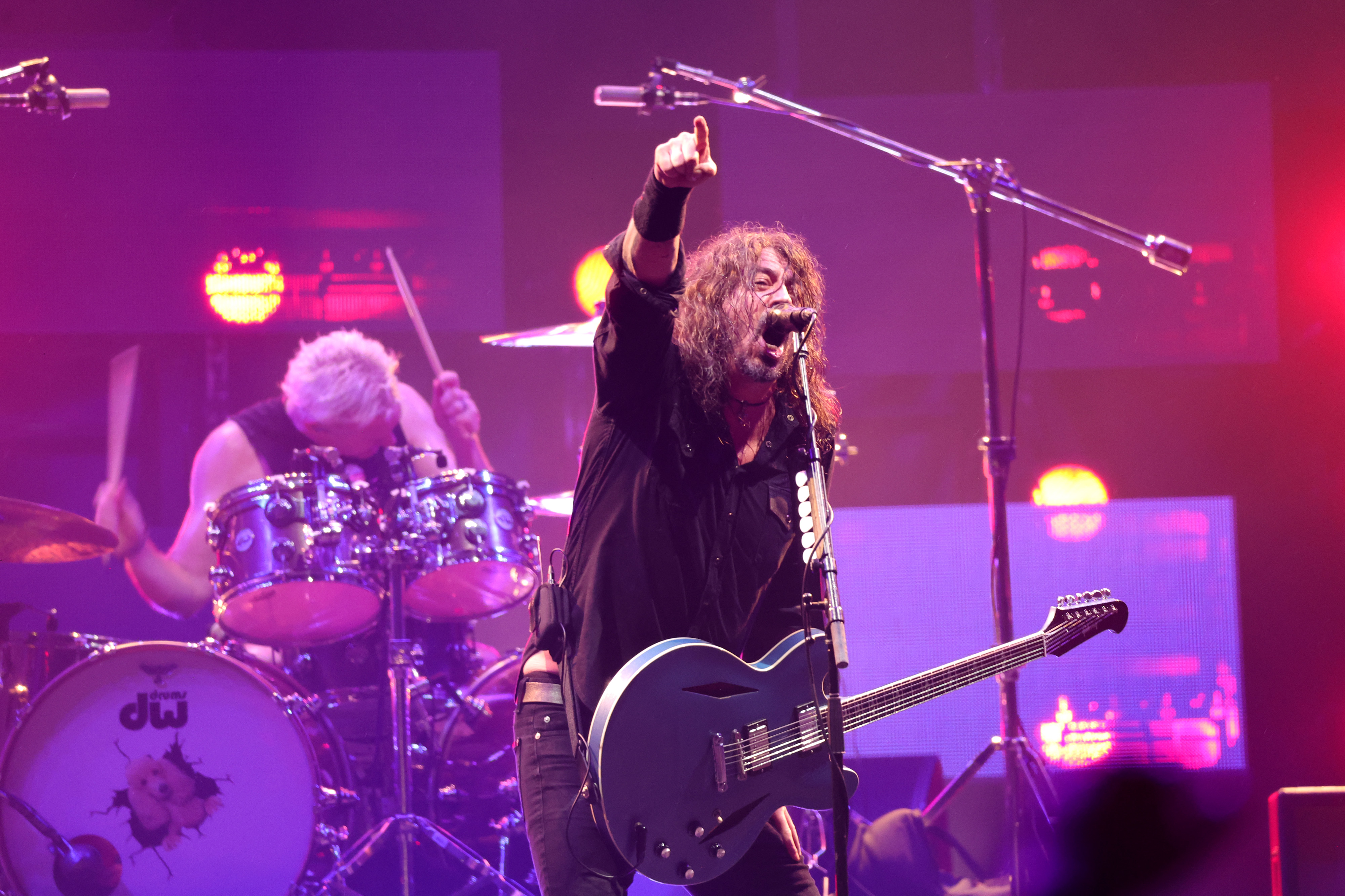 Dave Grohl performing energetically on stage with Taylor Hawkins playing drums in the background
