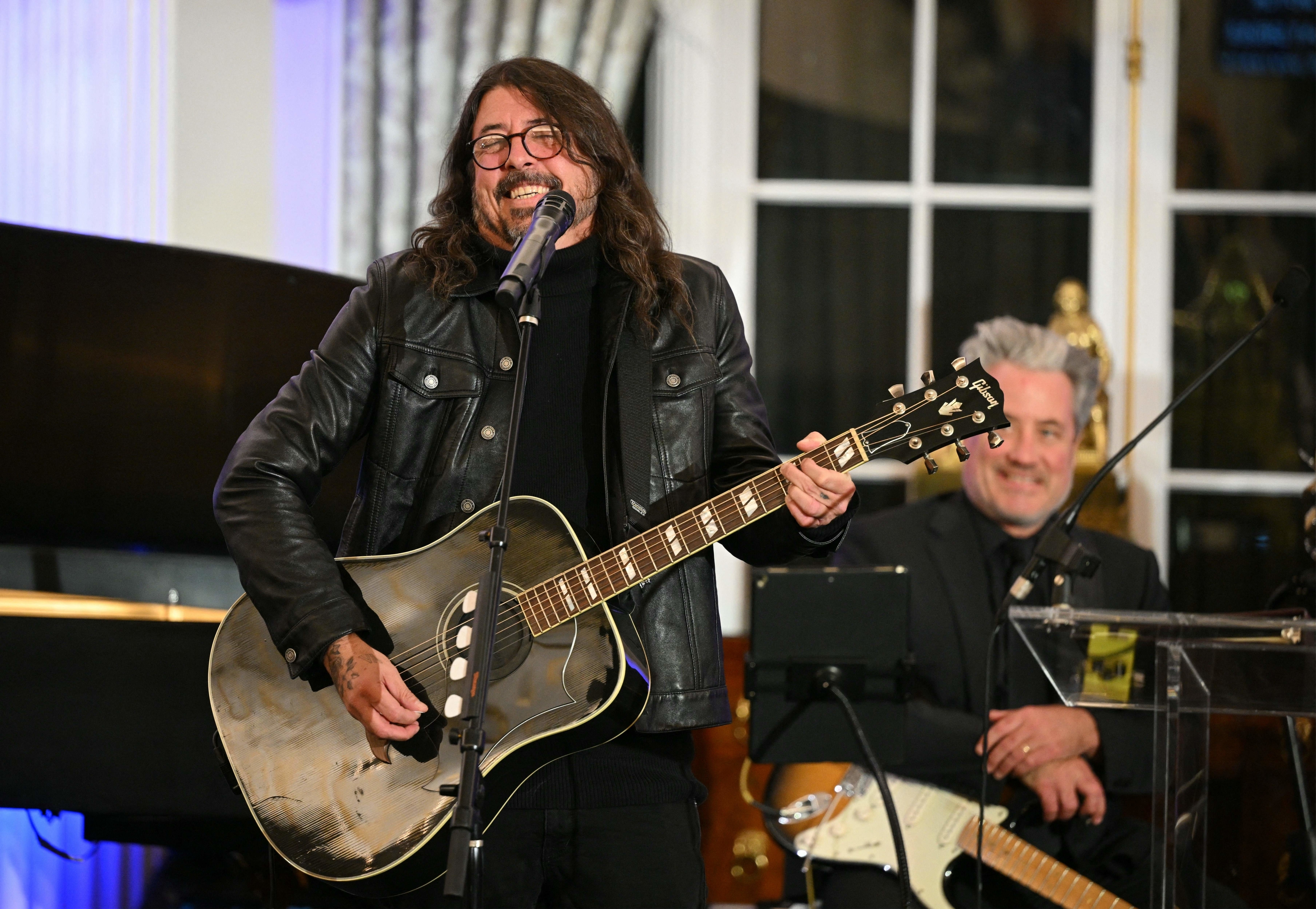 Dave Grohl plays an acoustic guitar and sings into a microphone on stage. A man in the background smiles while holding an electric guitar