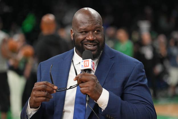 Shaquille O'Neal, dressed in a suit and tie, speaks into a microphone while holding glasses