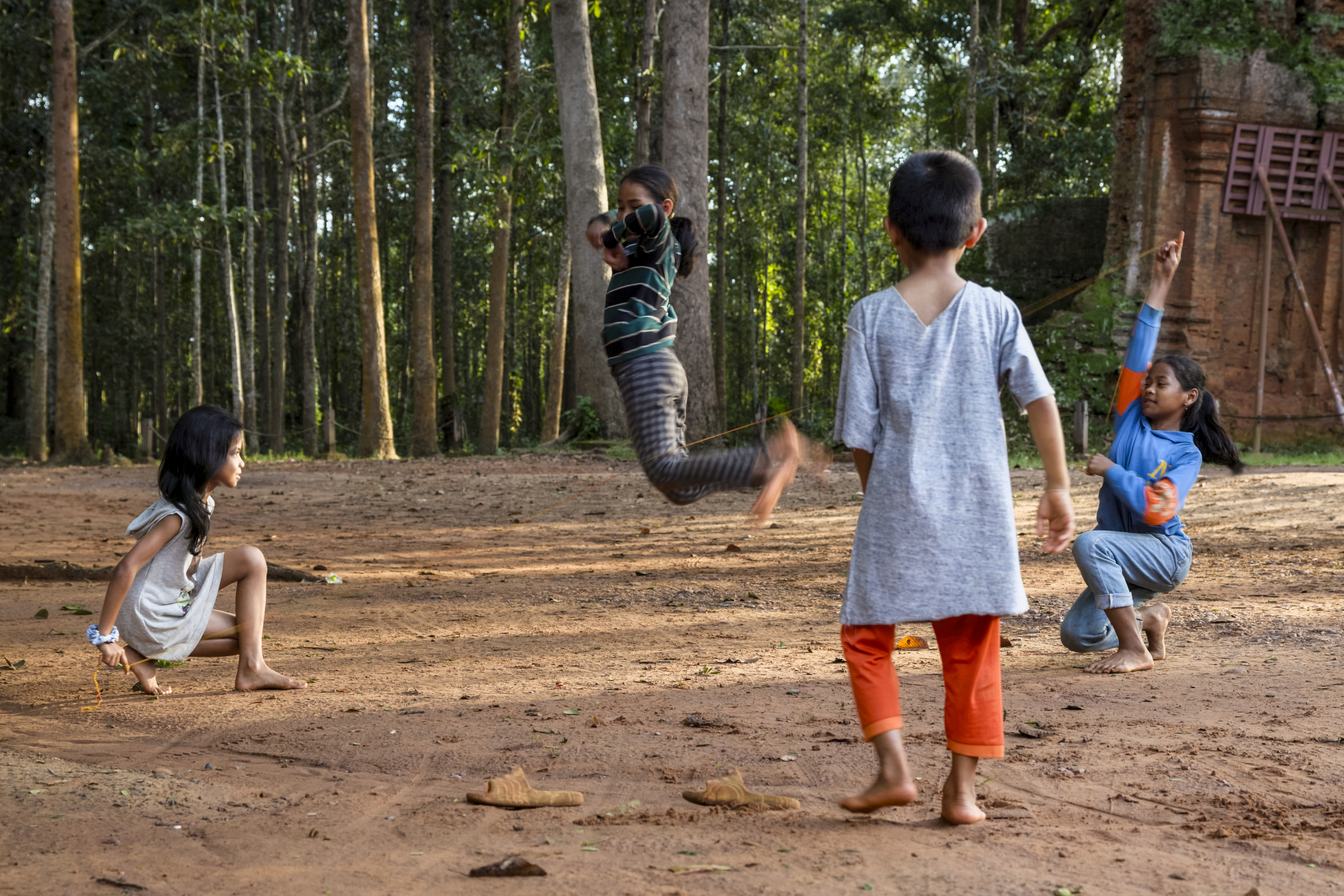 Four children are playing outside on dirt ground surrounded by trees. One child jumps in mid-air, while the others watch and play