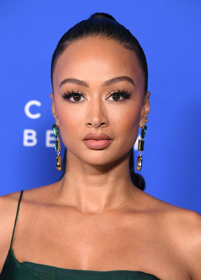Draya Michele is wearing long, emerald-green earrings and a matching, elegant dress, posing against a plain backdrop