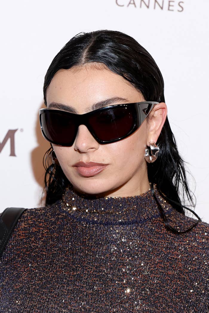 Charli XCX on the red carpet, wearing dark sunglasses, large silver hoop earrings, and a sparkling high-neck dress