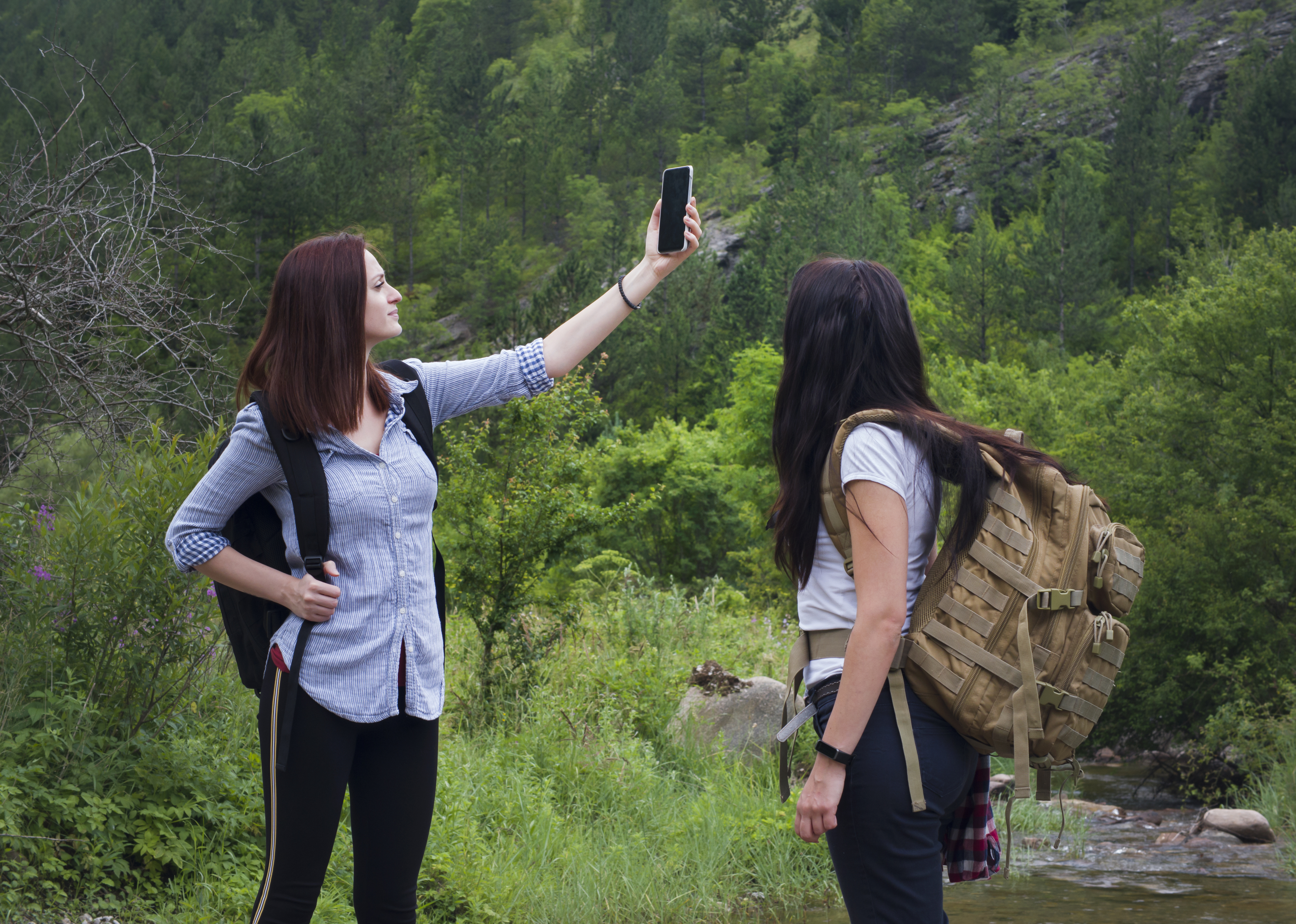 Two women are taking a selfie during a hike in a green, forested area. Both carry backpacks and appear to be enjoying the nature around them