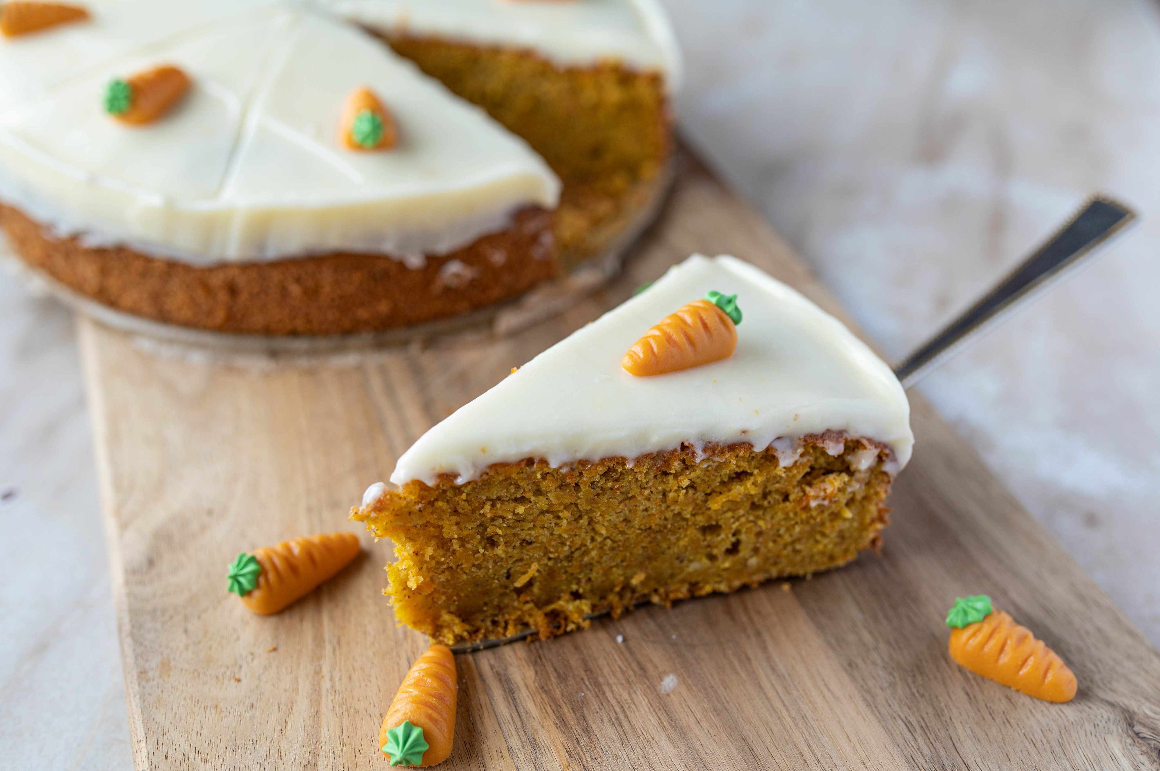 A slice of carrot cake with white icing and small carrot decorations, placed on a wooden board. The whole cake is visible in the background