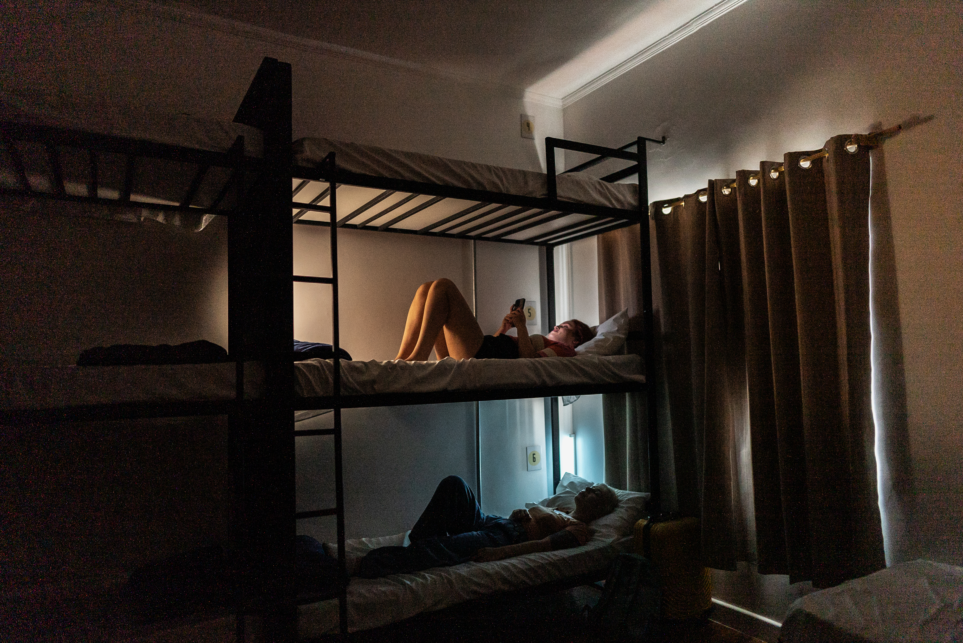 Two people rest on bunk beds in a dimly lit room. The person on the top bunk reads from a mobile device while laying down, and the person on the bottom bunk reclines