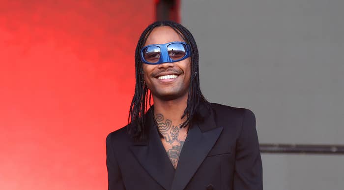 Steve Lacy on stage holding a microphone, wearing a black double-breasted coat and sunglasses, smiling