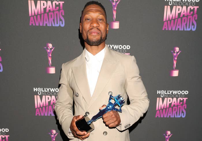 Man in tan suit holding a trophy, posing on the red carpet at the Hollywood Unlocked Impact Awards. Background features event logos repeated