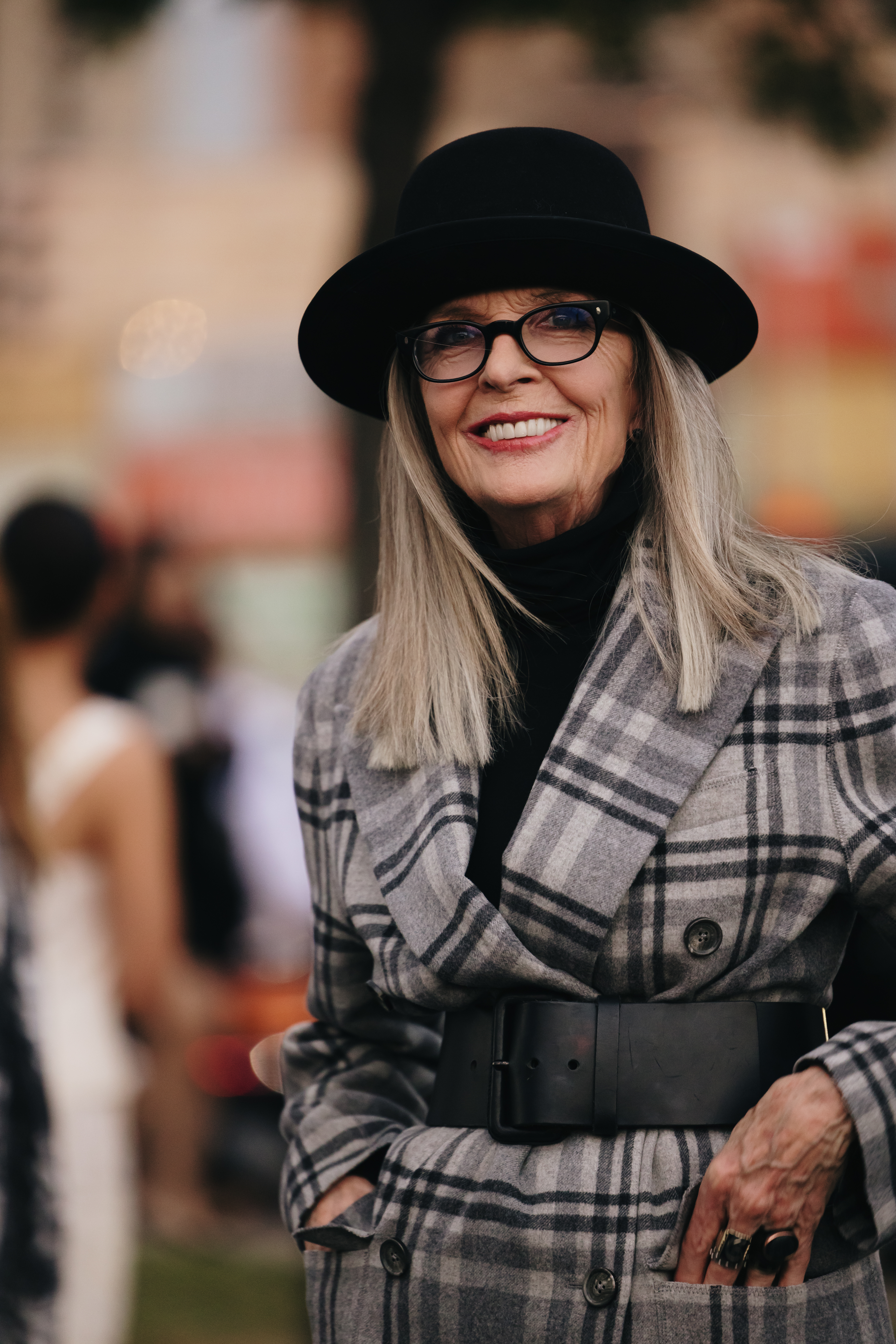 Diane Keaton smiles while wearing a plaid coat, black turtleneck, black wide-brim hat, and black belt at an outdoor event