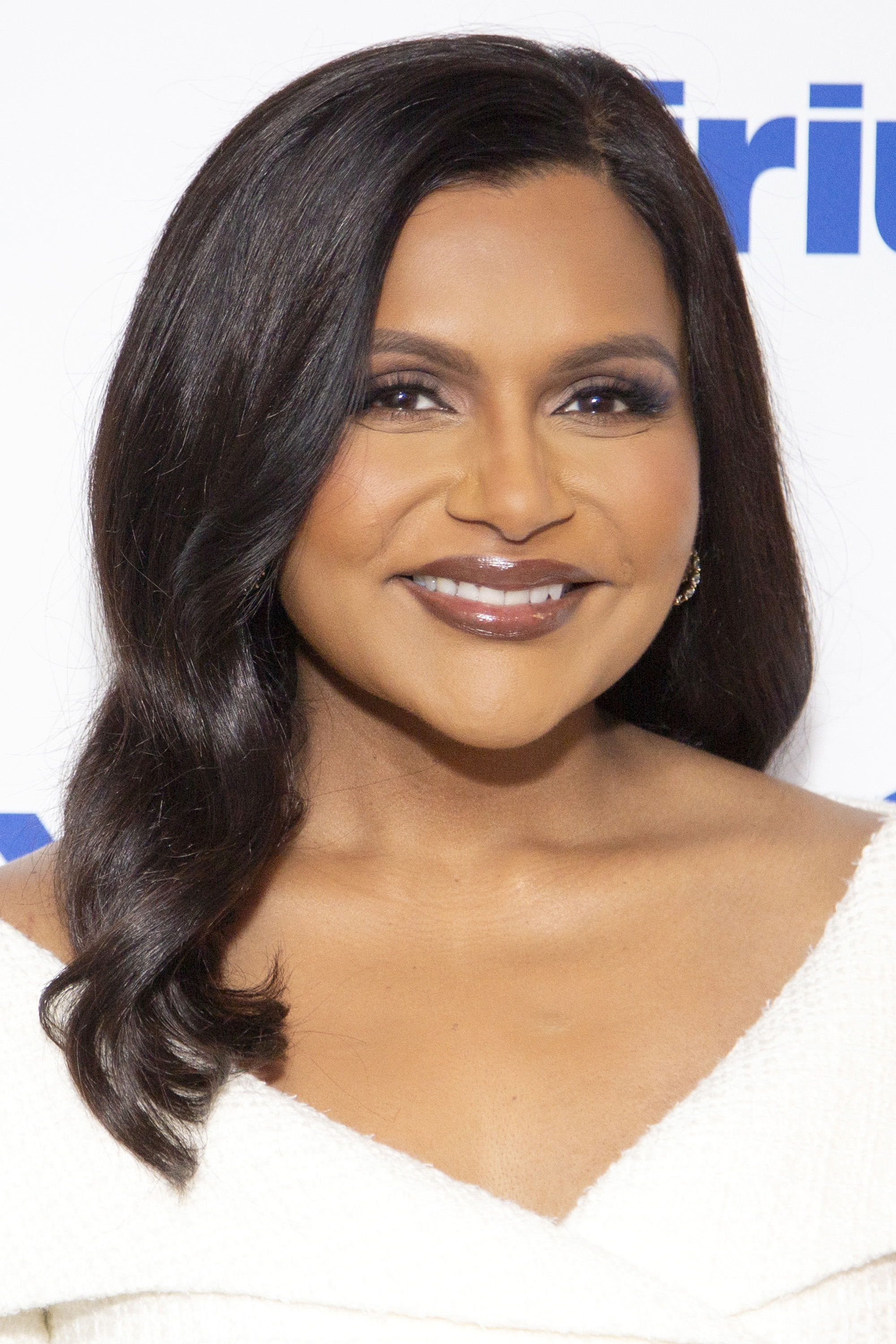Mindy Kaling smiles at an event, wearing an off-the-shoulder white dress