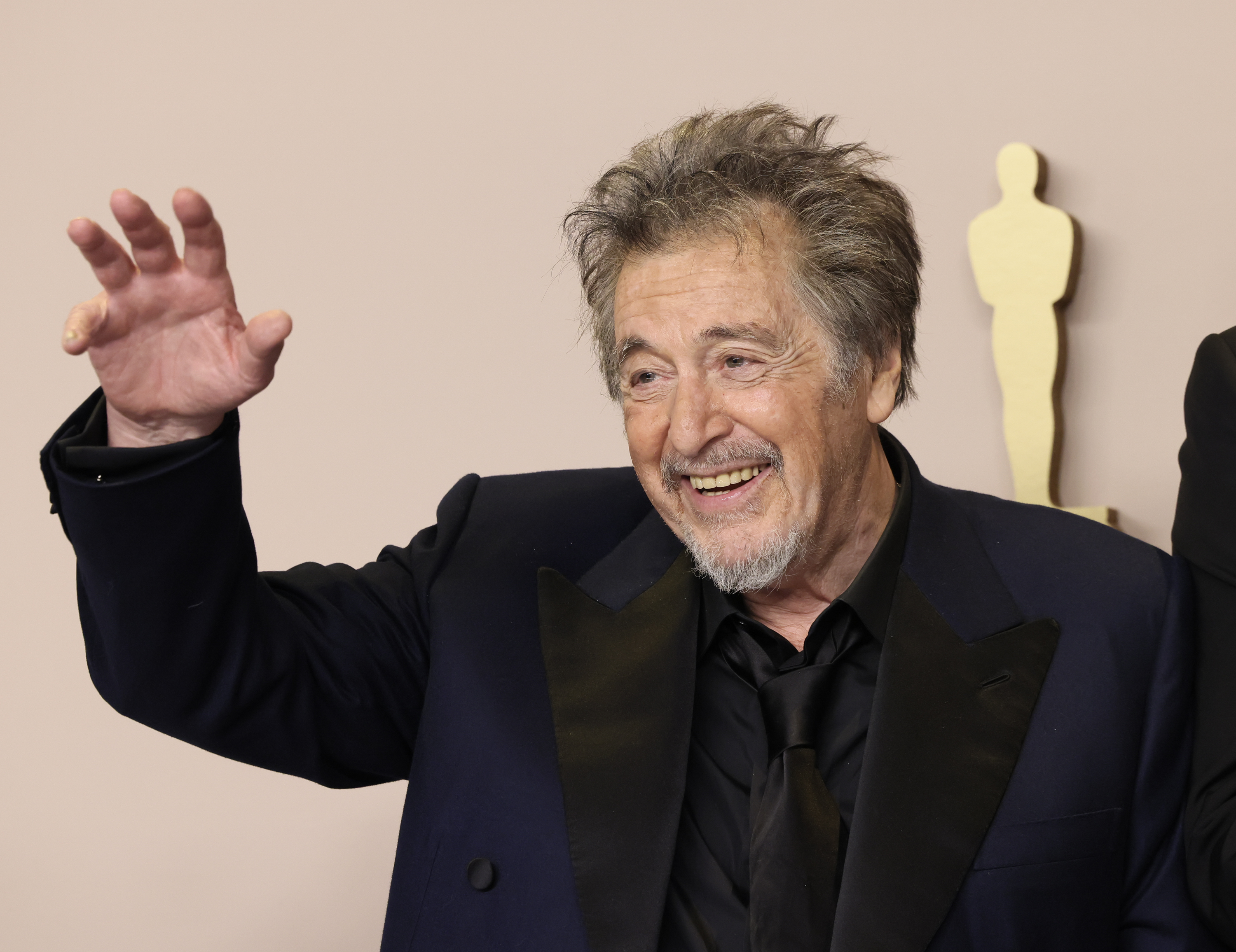 Al Pacino, wearing a dark suit and black shirt, smiles and waves at an event with an Oscar statue in the background
