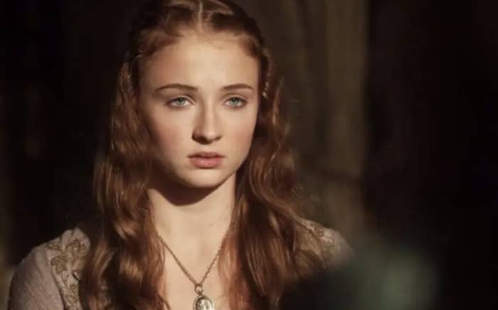 Sophie Turner as Sansa Stark, wearing a medieval-style dress and a pendant necklace, with a serious expression in a scene from &quot;Game of Thrones&quot;