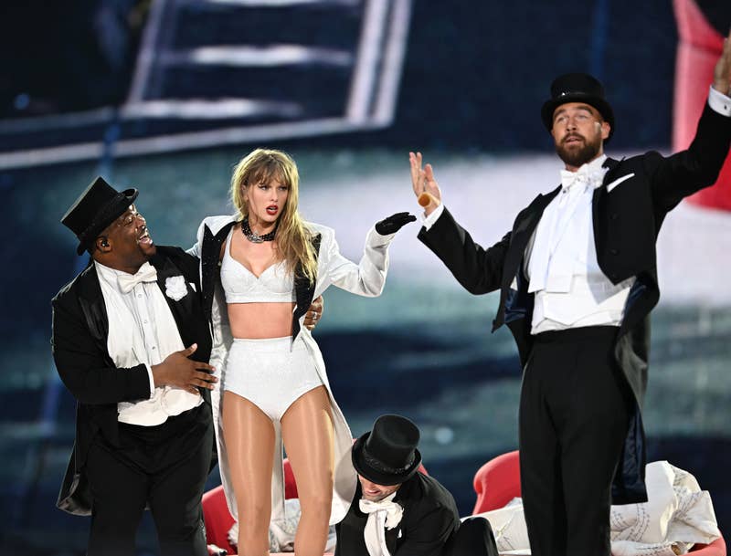 Taylor Swift is performing on stage with three backup dancers; she is wearing a white bodysuit and the dancers are dressed in tuxedos with top hats