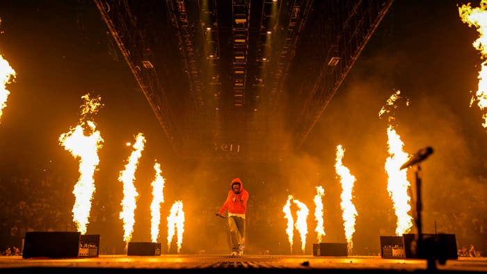 A performer on stage wearing a hoodie walks amid columns of fire during a live music concert, with a microphone in hand