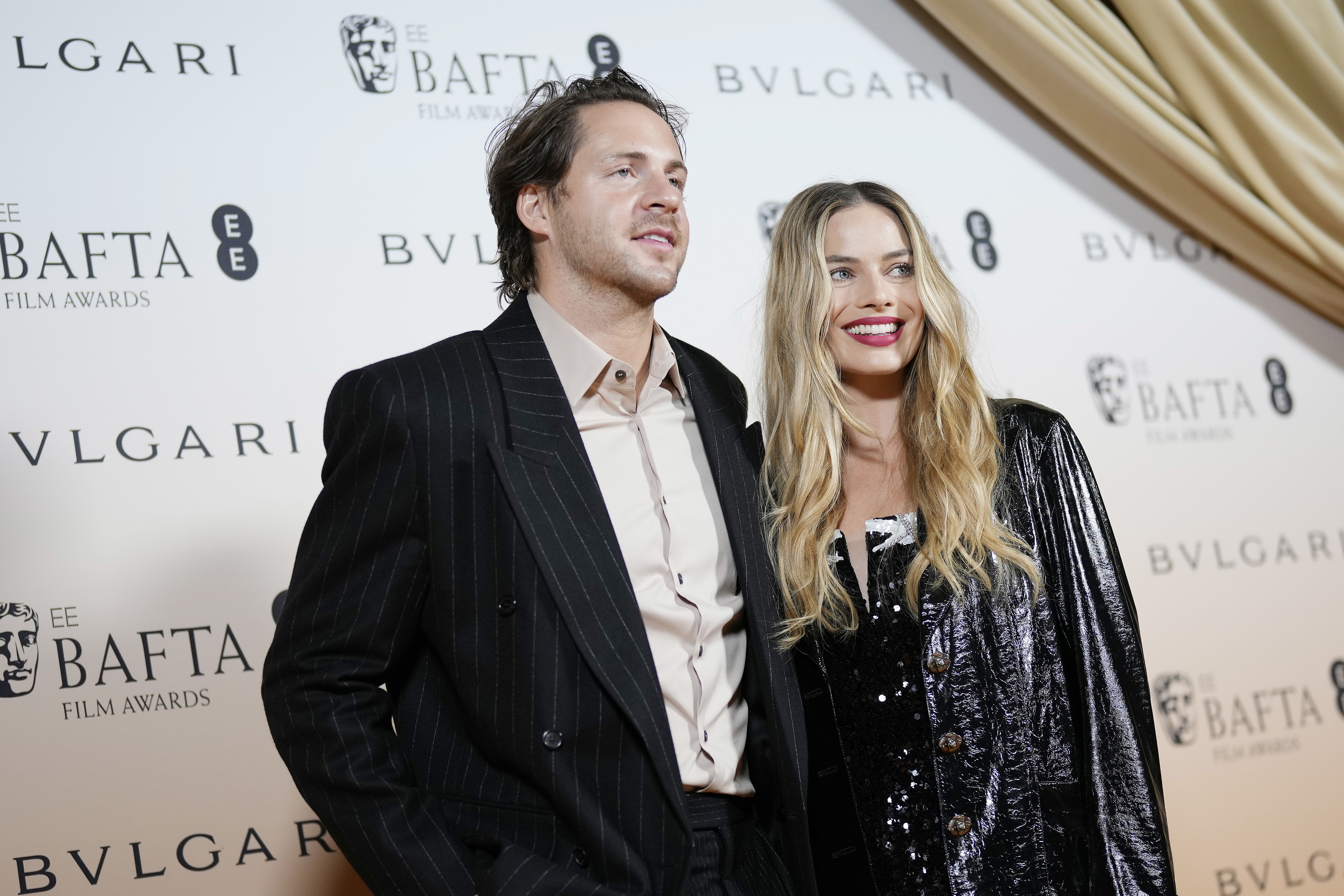 Tom Ackerley and Margot Robbie at the BAFTA Film Awards red carpet. Tom is wearing a suit, and Margot is in a sparkly outfit with a shiny jacket