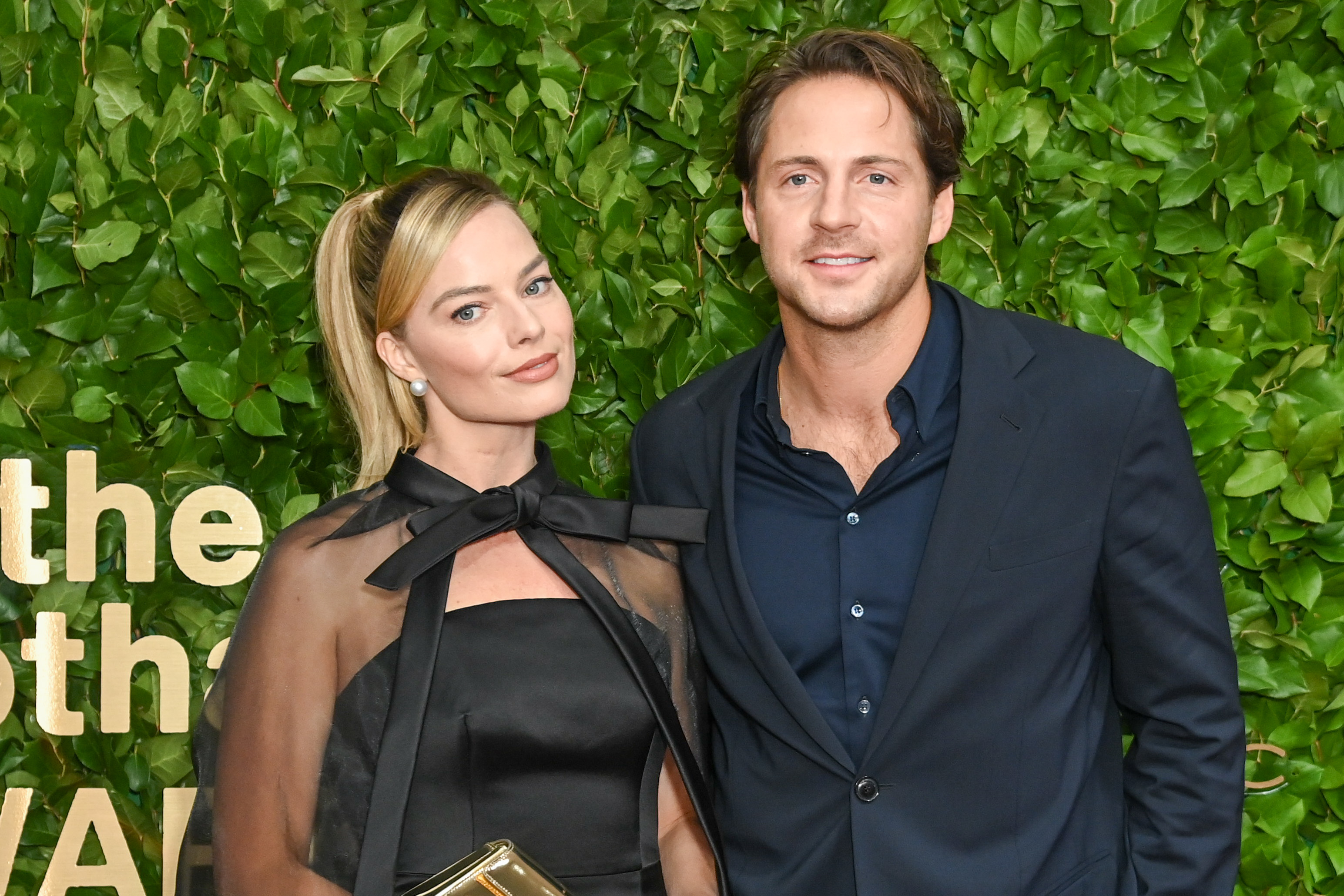 Margot Robbie, in a chic black dress with a sheer bow detail, posing with Tom Ackerley, in a dark suit against a greenery backdrop at a formal event