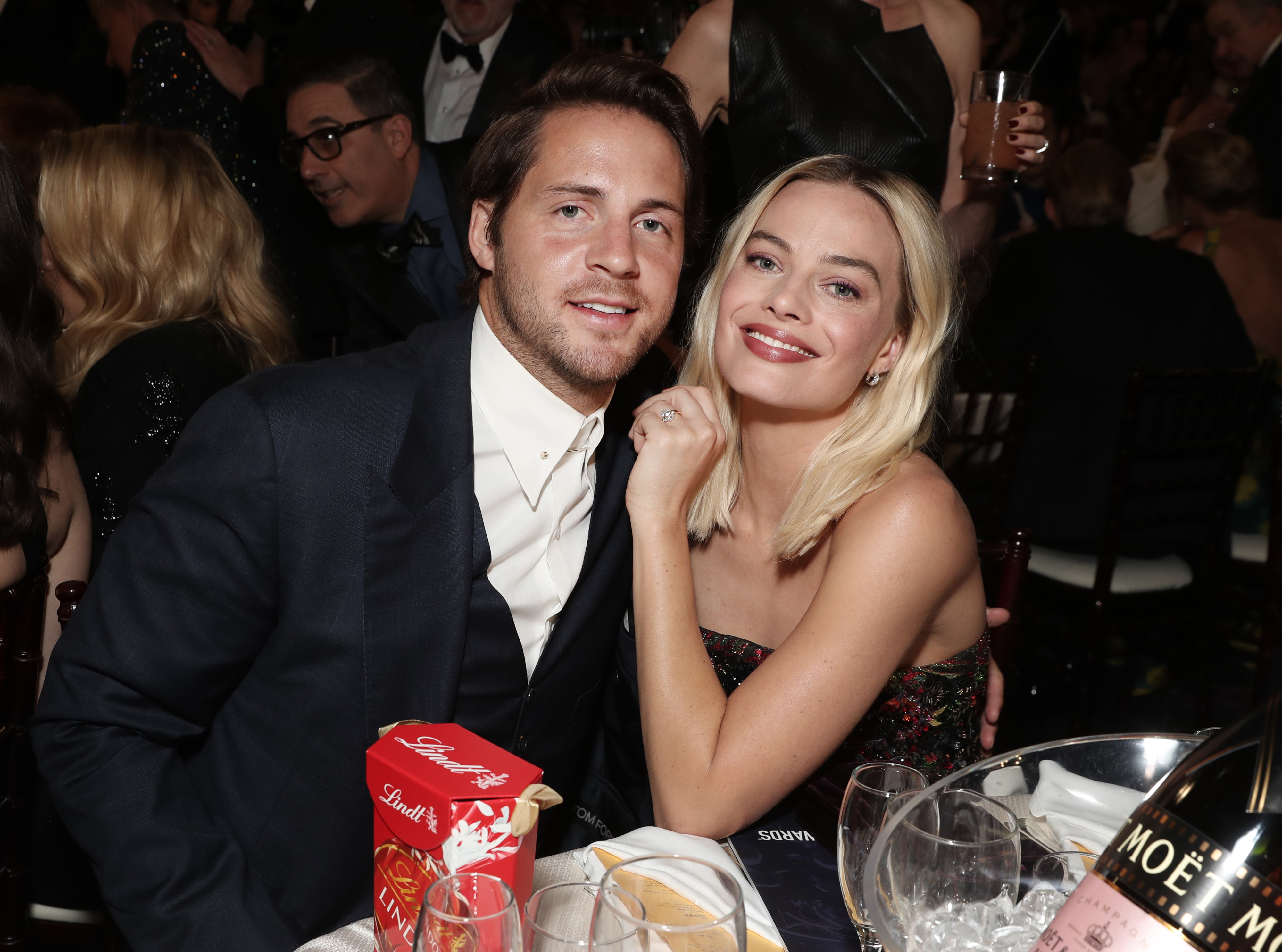 Margot Robbie in a strapless dress poses with Tom Ackerley at a formal event, with wine glasses and a Lindt chocolate box on the table