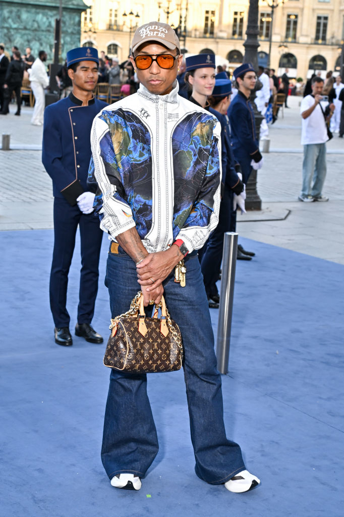 Pharrell Williams stands outdoors wearing a patterned jacket, jeans, and a cap. He carries a brown designer handbag and sunglasses while posing for the camera