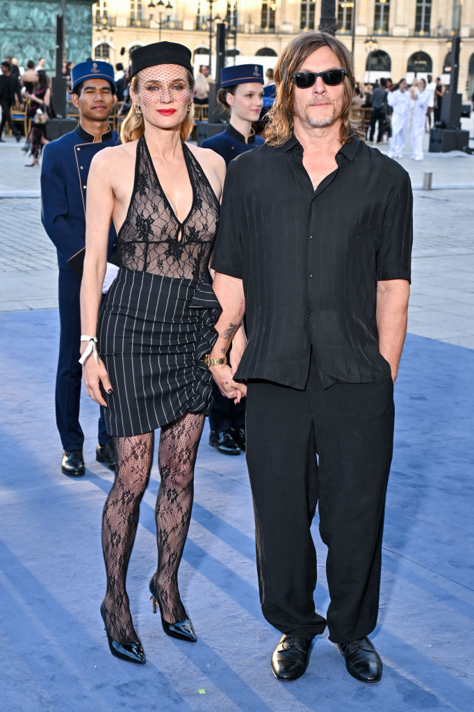 Diane Kruger in a lace top, pinstripe skirt, and fishnet stockings, and Norman Reedus in a black shirt and pants, stand together outdoors