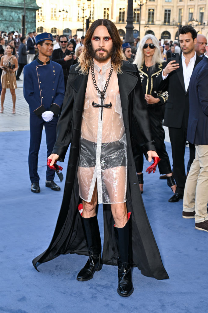 Jared Leto on the red carpet wearing a clear plastic outfit with a large black cross necklace and black boots, with a long black coat, surrounded by spectators