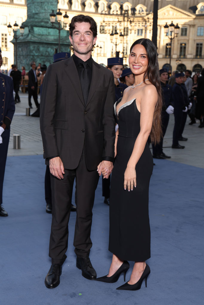 John Mulaney in a black suit and Olivia Munn in a black dress, holding hands and smiling at a formal event
