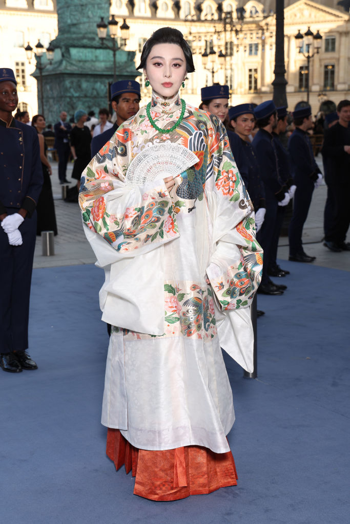 Fan Bingbing wearing a traditional, elegant embroidered outfit, holding a decorative fan, posing at an event with a blue carpet and people in the background
