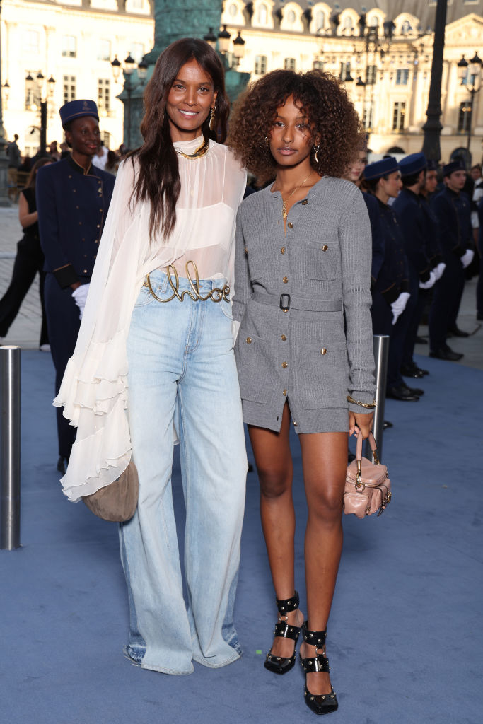 Liya Kebede and Raee Kebede pose together at an outdoor event. Liya wears flowy jeans with a sheer top, and Imaan wears a gray button-down dress with heels