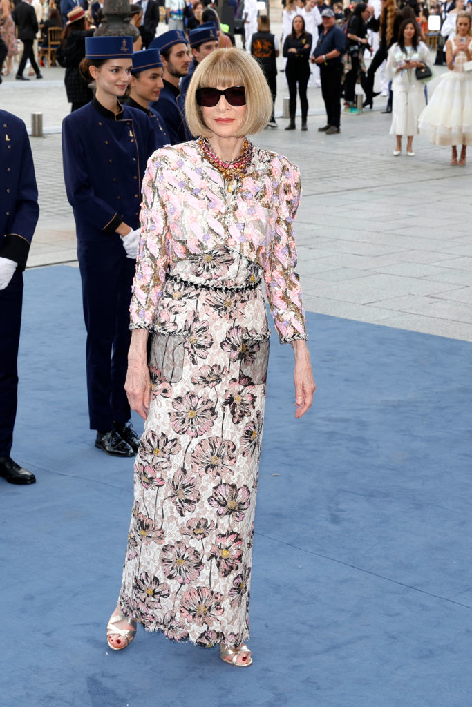 Anna Wintour on a blue carpet, wearing a floral-patterned dress with long sleeves, sunglasses, and a beaded necklace